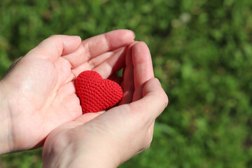 Red knitted heart in female palm hands on blurred nature background. Concept of love, health care,...