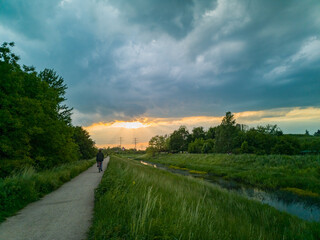 Fototapeta premium Beautiful cloudy sunset over long dark river in park with biker on path