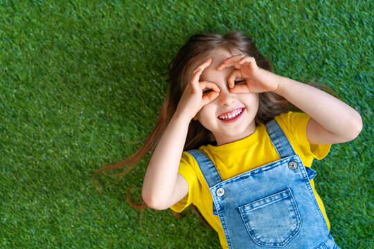 cheerful little girl in a denim jumpsuit, lies on a green carpet, lawn.