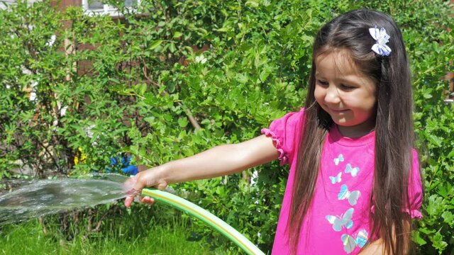 Little cute girl holds a watering hose in her hands. A child watering plants in the garden. The concept of childhood and motherhood