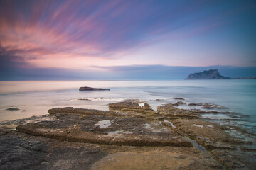 Paisaje costero al atardecer con el Peñon de Ifach en el horizonte. El mar está en calma y el cielo nublado.