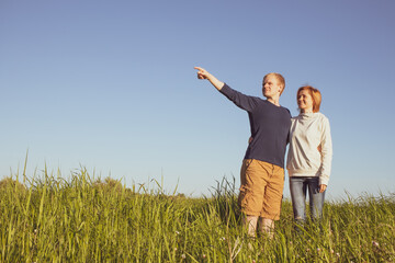 Young happy couple, man and woman, stand in the grass and look to the side. The man shows hand into the distance. Big place for text on sky background. Sunny weather. The concept of optimistic view.