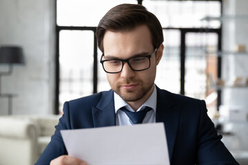 Close up of serious male employer in glasses read consider paperwork letter or correspondence in office, concentrated Caucasian young businessman analyze paper document or report at workplace
