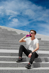 Young sporty man sitting on stairs outdoors drinking water from eco plastic bottle against blue sky background . Resting man in sunglasses. Concept of sport, activity, outdoor active life style.