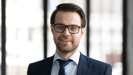 Fototapeta premium Close up headshot portrait of smiling young Caucasian businessman in formal suit and glasses pose in office, happy successful confident male leader in spectacles show power and motivation at workplace