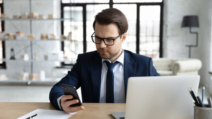 Fototapeta na wymiar Serious young Caucasian businessman sit at office desk multitask work on laptop and smartphone, focused male employer text message on cellphone gadget, use computer, new technology concept