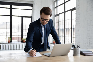Concentrated young Caucasian businessman in formal suit glasses busy working on laptop making...