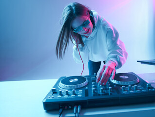 Cool young girl DJ mixes music on a mixing console and laptop, in stylish clothes, glasses on a...