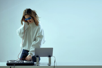 Cool young girl DJ mixes music on a mixing console and laptop, in stylish clothes, glasses on a...