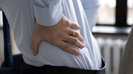 Crop close up of male employee touch lower back suffering from muscular spasm or strain sit in incorrect posture, unwell worker struggle with backache, have painful feeling, sedentary life concept