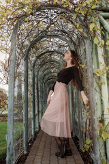 Beautiful girl in the arch with plants. Young woman in a pink skirt and black sweater on a walk in the park.