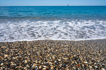 Foamy wave on calm beach. Shore with pebbles. Blue sky seascape with sailboat on the horizon.