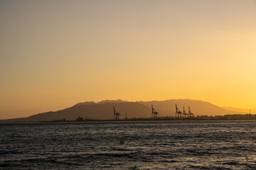 Summer sunset. Orange sky over the sea, mountains on the horizon and port container cranes.