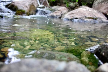 Mountain river - a small waterfall on a river with crystal clear water that flows among gray stones in a green forest on a cloudy summer day. Big stones near the pond