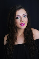 Naklejka premium beautiful brunette young girl happily smiling, looking at camera with fresh make up on black background, pink lipstick curly hair over shoulders, in black dress studio shot, vertical image