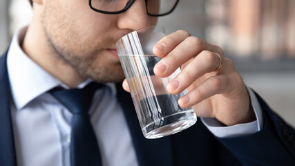 Close up of young businessman drink pure crystal mineral water from glass feeling dehydrated at workplace, thirsty male employee enjoy clean clear aqua, refresh body, healthy lifestyle concept