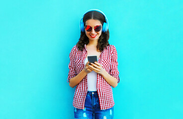 Portrait of young smiling woman with smartphone in wireless headphones listening to music wearing a pink sunglasses on colorful blue background