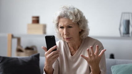 Angry mature woman sit on couch at home feel frustrated by slow internet connection on cellphone,...