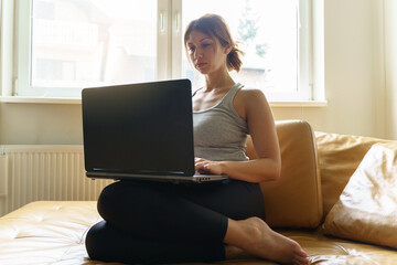 Fototapeta premium Young adult caucasian woman working on the laptop computer from home sitting on the sofa bed in day taping online female freelancer or shopping online efficiency and expertise making money concept
