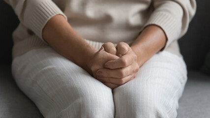 Close up of upset mature woman sit on sofa hold hands joined clenched on laps feel worried at home alone, pensive elderly female anxious about decision, thinking or pondering, elderly solitude concept