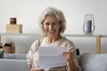 Smiling elderly 60s woman sit on couch in living room feel overjoyed reading good news in postal...