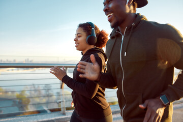 Morning sport. Young smiling african couple in headphones jogging together on the bridge. Happy African man and woman enjoying running outdoors