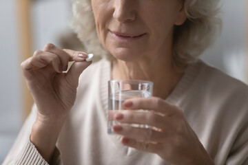 Close up of mature woman having antibiotic or aspirin pill drug with glass of water at home, unhealthy senior female take daily dose of vitamins or supplements, elderly healthcare, medicine concept