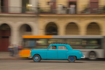 Fototapeta na wymiar Panning shot of classic blue car with blurred bus and building in background. Sunrise shot in Havana, Cuba