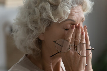 Close up of tired elderly woman rub massage eyes take off glasses suffering from headache or migraine, exhausted mature 60s female have dizziness or blurry vision form high blood pressure