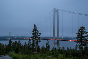 Obraz premium Picture of the illuminated Höga Kustenbron, a suspension bridge crossing the river Ångermanälven near Veda, Sweden taken on a late evening with low cloud cover around midsummer. 