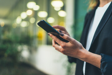Closeup photo business woman wearing suit, looking smartphone  in hands. Open space loft office. Panoramic windows background. Wide mockup. Film effect