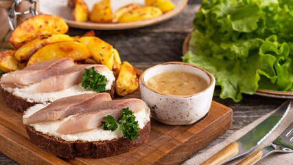 Sandwich with herring fillets, baked potato and sauce on wooden board.