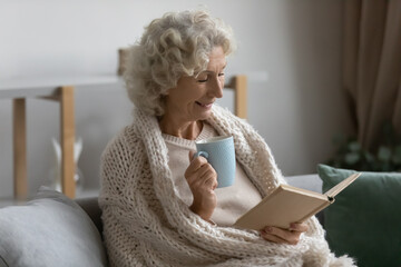 Smiling mature 50s woman rest on sofa in living room, drink tea read book, happy senior 60s...