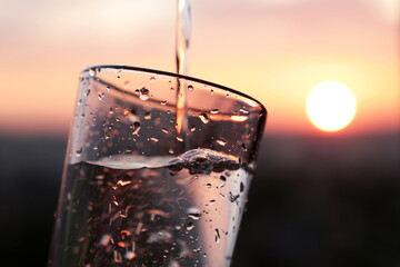 Pouring clean water into drinking glass on sunset background. Concept of health and freshness,...