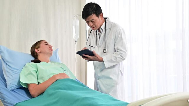 4K Senior doctor talks to caucasian patient on bedside in the hospital as he holds a tablet computer