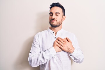 Young handsome man with beard wearing casual t-shirt standing over white background smiling with hands on chest, eyes closed with grateful gesture on face. Health concept.