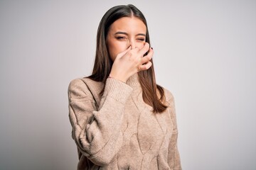 Young beautiful brunette woman wearing casual sweater standing over white background smelling...