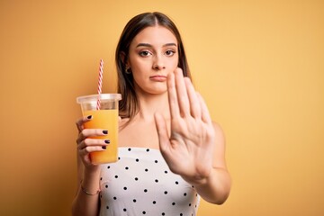 Young beautiful brunette woman drinking healthy orange juice over yellow background with open hand doing stop sign with serious and confident expression, defense gesture