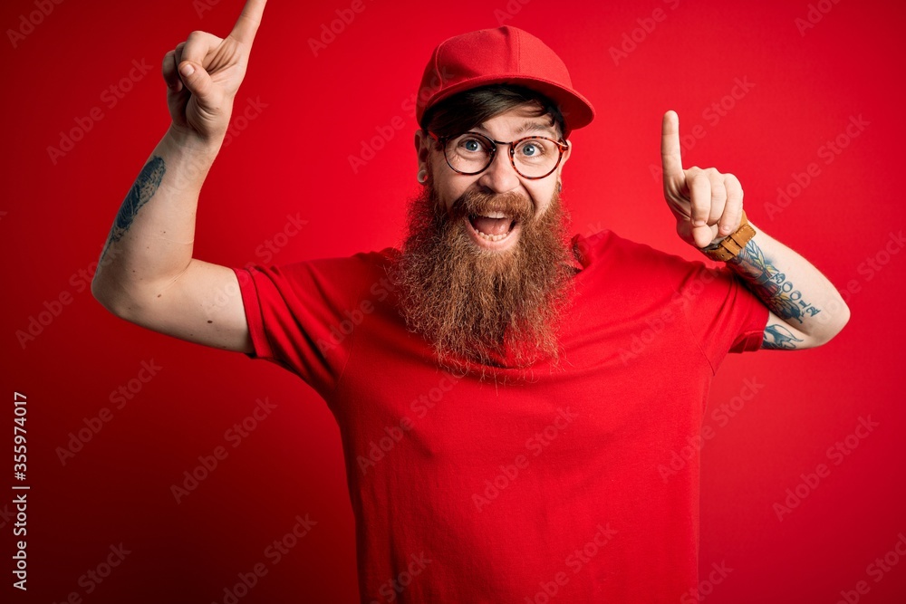 Sticker Young handsome delivery man wearing glasses and red cap over isolated background smiling amazed and surprised and pointing up with fingers and raised arms.