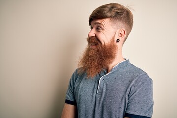 Handsome Irish redhead man with beard and arm tattoo standing over isolated background looking away...