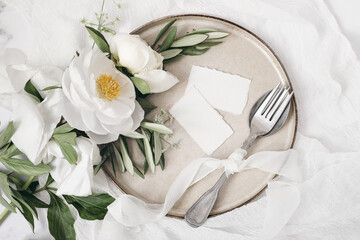 Festive summer wedding scene. Marble table setting with cutlery, olive branches, white peony flowers, stoneware plate and silk ribbon. Blank place cards mockups. Restaurant menu concept. Flat lay, top