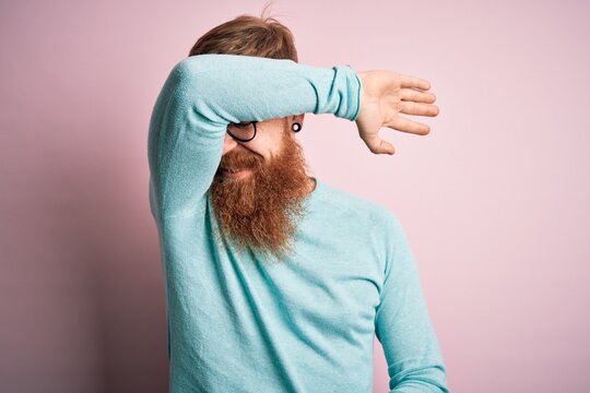 Handsome Irish redhead man with beard wearing glasses over pink isolated background covering eyes with arm smiling cheerful and funny. Blind concept.