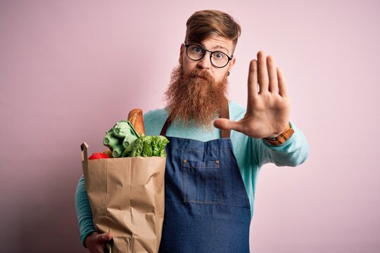 Redhead Irish Supermarket Worker Man With Beard Wearing Apron And Holding Fresh Groceries With Open Hand Doing Stop Sign With Serious And Confident Expression, Defense Gesture