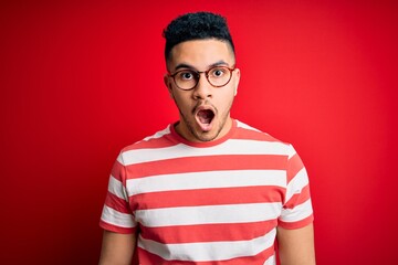Young handsome man wearing casual striped t-shirt and glasses over isolated red background afraid and shocked with surprise and amazed expression, fear and excited face.