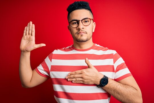 Young handsome man wearing casual striped t-shirt and glasses over isolated red background Swearing with hand on chest and open palm, making a loyalty promise oath