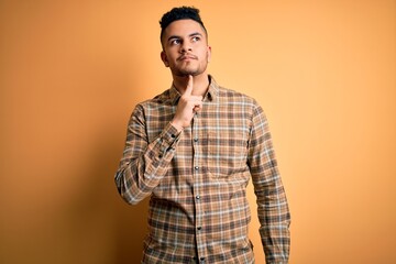 Young handsome man wearing casual shirt standing over isolated yellow background Thinking concentrated about doubt with finger on chin and looking up wondering
