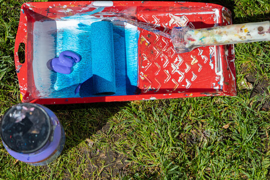 On the grass is a can of paint and a tray. The tray has blue paint and a paint roller.
Concept: preparation for painting.