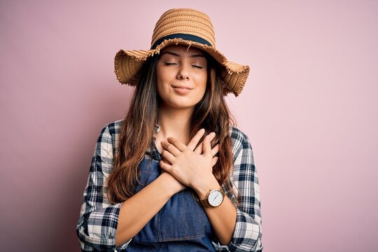 Young beautiful brunette farmer woman wearing apron and hat over pink background smiling with hands on chest with closed eyes and grateful gesture on face. Health concept.
