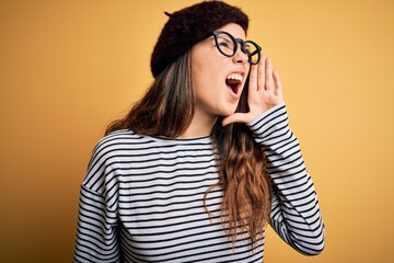 Young beautiful brunette woman wearing french beret and glasses over yellow background shouting and screaming loud to side with hand on mouth. Communication concept.