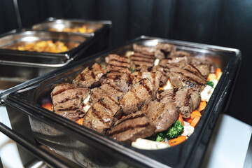 Stainless hotel pans on food warmers with various meals. Grilled beef steaks laying on vegetable....